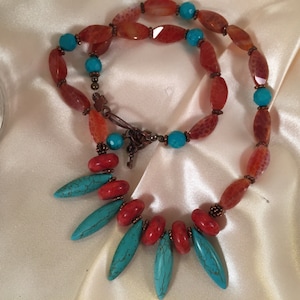 May include: A layered necklace with orange and turquoise beads. The top layer is a long strand of oval-shaped orange beads with turquoise accents. The bottom layer is a shorter strand of orange beads with turquoise accents and five turquoise teardrop-shaped beads.