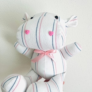 May include: A white and pink striped stuffed hippopotamus with pink heart-shaped buttons for eyes and a pink bow. The hippopotamus is embroidered with the text "2022 Dara".