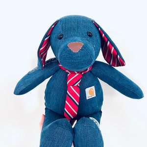 May include: A blue denim dog plush toy wearing a red and white striped tie. The toy has a brown nose and black eyes. The toy has a Carhartt logo on the chest.