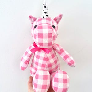 May include: A stuffed unicorn toy with a pink and white gingham pattern. The unicorn has a white horn with black heart accents, a pink bow, and black button eyes. The toy is soft and plush.
