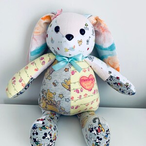 K&ouml;nnte beinhalten: Ein handgemachtes Patchwork-Hasenspielzeug mit einer rosa Schleife auf dem Kopf. Der Hase ist aus verschiedenen Stoffen gefertigt, darunter ein Winnie-the-Pooh-Druck, ein Mickey-Mouse-Druck und ein Blumendruck. Der Hase hat einen herzf&ouml;rmigen Aufn&auml;her auf der Brust, auf dem "Mommy's Sweet Heart" steht.