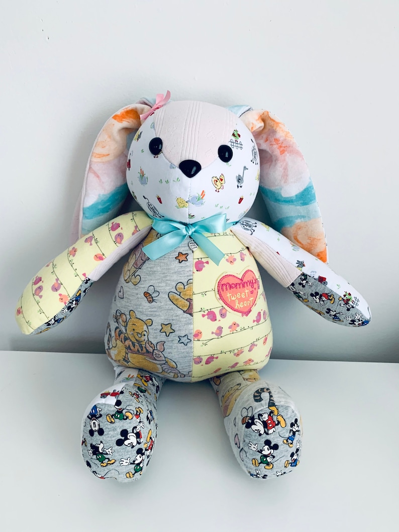 K&ouml;nnte beinhalten: Ein handgemachtes Patchwork-Hasenspielzeug mit einer rosa Schleife auf dem Kopf. Der Hase ist aus verschiedenen Stoffen gefertigt, darunter ein Winnie-the-Pooh-Druck, ein Mickey-Mouse-Druck und ein Blumendruck. Der Hase hat einen herzf&ouml;rmigen Aufn&auml;her auf der Brust, auf dem "Mommy's Sweet Heart" steht.