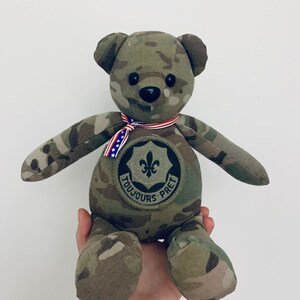 May include: Camouflage teddy bear with a black nose and black eyes. The bear is wearing a red, white, and blue ribbon and a patch that says "Toujours Pret" with a fleur-de-lis.