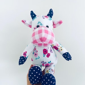 May include: A white plush cow toy with pink and white gingham fabric on the face, floral fabric on the body, and black polka dot fabric on the hooves and ears.