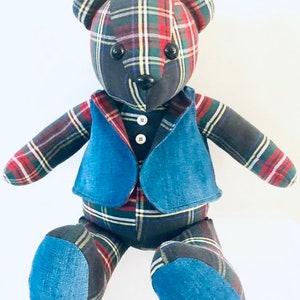 May include: A plaid teddy bear wearing a denim vest with three buttons. The bear has black eyes and a black nose.