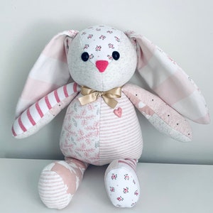 K&ouml;nnte beinhalten: Ein handgemachtes Patchwork-Hasenspielzeug aus rosa-wei&szlig; gestreiftem Stoff, Blumenmustern und einem rosa Herz-Knopf. Der Hase hat ein goldenes Band um den Hals.