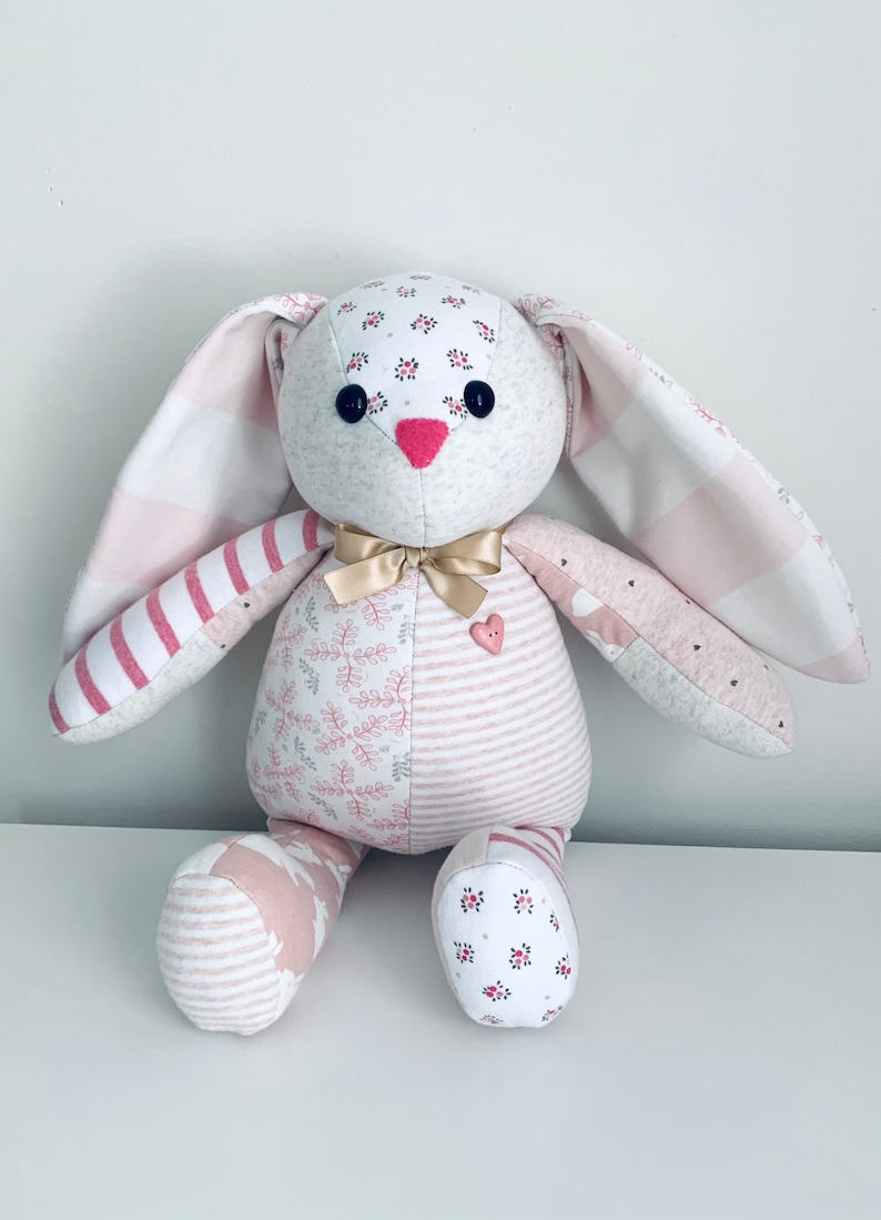 K&ouml;nnte beinhalten: Ein handgemachtes Patchwork-Hasenspielzeug aus rosa-wei&szlig; gestreiftem Stoff, Blumenmustern und einem rosa Herz-Knopf. Der Hase hat ein goldenes Band um den Hals.