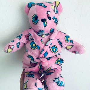 May include: A pink teddy bear wearing a matching pink robe. The bear and robe are covered in a pattern of donuts, ice cream cones, and candy. The bear has black eyes and a black nose. The robe has a soft, plush texture.
