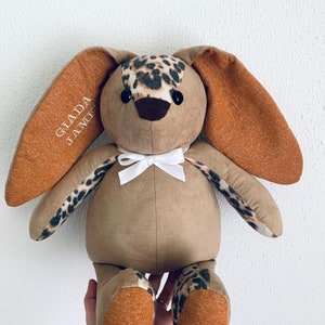 K&ouml;nnte beinhalten: Ein brauner Pl&uuml;schhase mit einer wei&szlig;en Schleife, braunen Ohren mit orangenen Spitzen und einem Leopardenmuster auf dem K&ouml;rper. Der Hase hat schwarze Knopfaugen und der Text "GLADA JAMIE" ist auf ein Ohr gestickt.