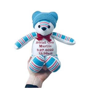 May include: A white plush teddy bear wearing a blue hat with pink and blue striped fabric. The bear is embroidered with the name "Adriel Cruz Murillo", the date "9.27.2022", and the time "12:36am".