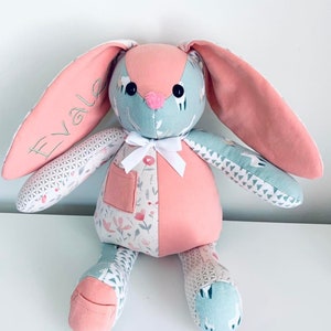 K&ouml;nnte beinhalten: Ein rosa-blauer Patchwork-Hase mit wei&szlig;en Stoffohren. Der Hase hat eine wei&szlig;e Schleife und eine Tasche auf der Brust. Der Hase ist mit dem Namen "Evale" bestickt.