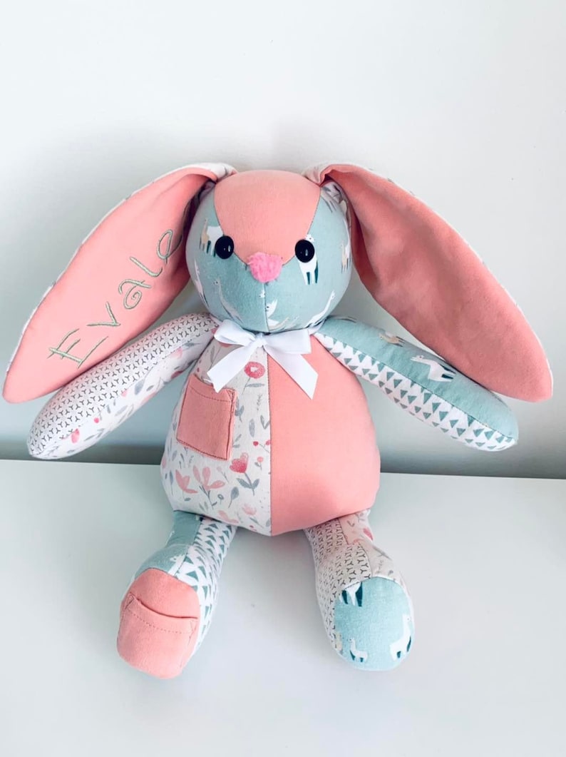 K&ouml;nnte beinhalten: Ein rosa-blauer Patchwork-Hase mit wei&szlig;en Stoffohren. Der Hase hat eine wei&szlig;e Schleife und eine Tasche auf der Brust. Der Hase ist mit dem Namen "Evale" bestickt.
