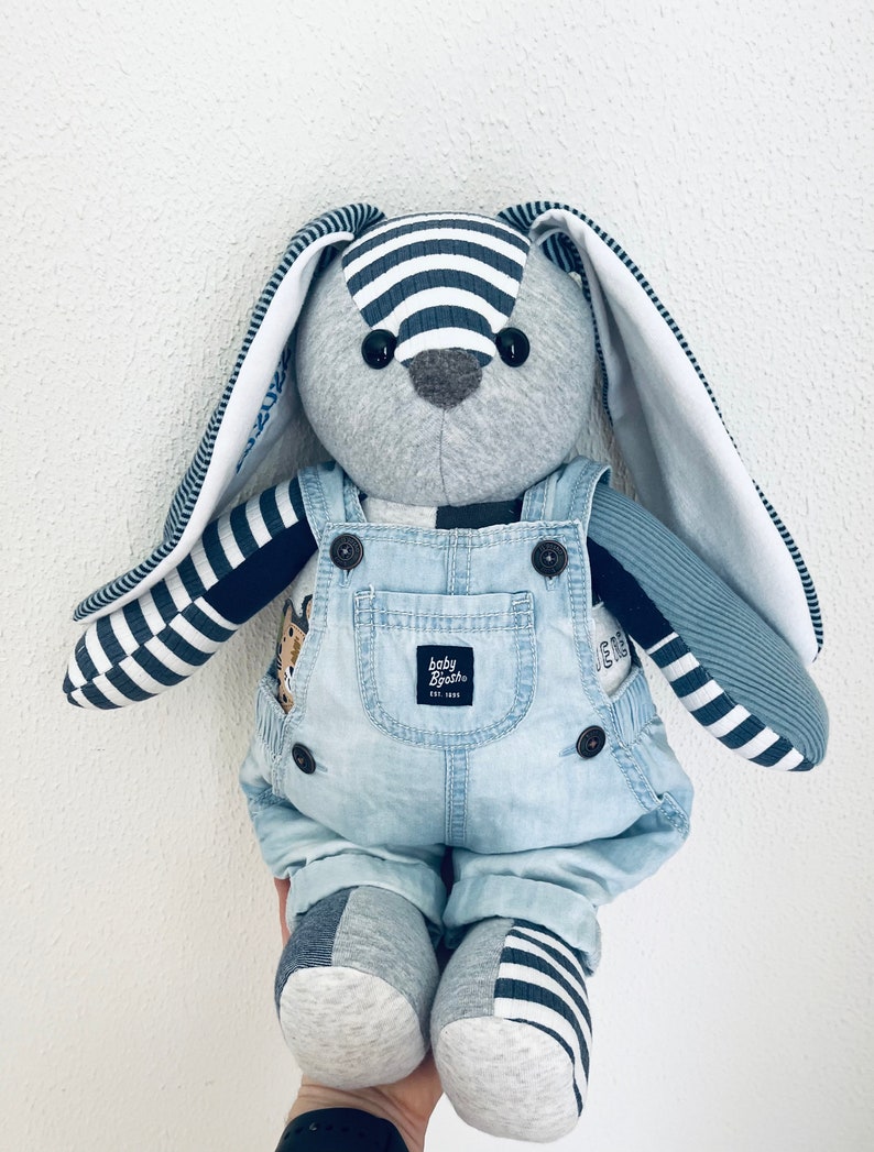 K&ouml;nnte beinhalten: Ein graues und blaues Stofftier-Kaninchen, das eine Jeanslatzhose mit dem Logo "baby bash" tr&auml;gt. Das Kaninchen hat lange, wei&szlig;e Ohren mit schwarz-wei&szlig; gestreiftem Besatz.