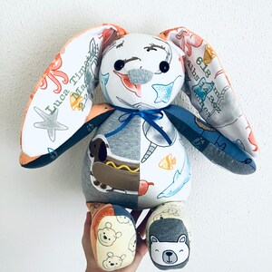 K&ouml;nnte beinhalten: Ein handgemachtes Pl&uuml;sch-Hasenspielzeug aus wiederverwendeten Babykleidern. Der Hase hat ein graues Gesicht mit schwarzen Augen und ein blaues Band um den Hals. Die Ohren des Hasen sind aus einem Stoff mit einem Seepferdchen- und Seesternmuster. Der K&ouml;rper des Hasen ist aus verschiedenen Stoffen gefertigt, darunter ein grauer Stoff mit einem Hotdog-Muster, ein wei&szlig;er Stoff mit einem Fischmuster und ein blauer Stoff mit einem Walmuster. Die F&uuml;&szlig;e des Hasen sind aus einem gelben Stoff mit einem B&auml;renmuster.