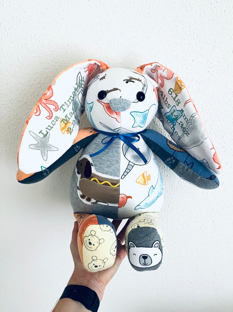 K&ouml;nnte beinhalten: Ein handgemachtes Pl&uuml;sch-Hasenspielzeug aus wiederverwendeten Babykleidern. Der Hase hat ein graues Gesicht mit schwarzen Augen und ein blaues Band um den Hals. Die Ohren des Hasen sind aus einem Stoff mit einem Seepferdchen- und Seesternmuster. Der K&ouml;rper des Hasen ist aus verschiedenen Stoffen gefertigt, darunter ein grauer Stoff mit einem Hotdog-Muster, ein wei&szlig;er Stoff mit einem Fischmuster und ein blauer Stoff mit einem Walmuster. Die F&uuml;&szlig;e des Hasen sind aus einem gelben Stoff mit einem B&auml;renmuster.