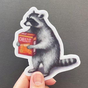 May include: A raccoon holding a box of Cheez-It crackers. The box is red and orange with the words "Cheez-It" in white letters. The raccoon is standing on its hind legs and has a black and white striped tail.