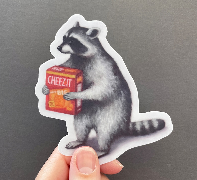 May include: A raccoon holding a box of Cheez-It crackers. The box is red and orange with the words "Cheez-It" in white letters. The raccoon is standing on its hind legs and has a black and white striped tail.