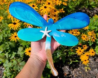 May include: A blue and gold dragonfly wall decor with a white seashell accent. The dragonfly is made of resin and has a glittery finish.
