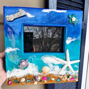May include: A blue, green, and brown resin picture frame with a beach scene. The frame features white foam waves, seashells, starfish, and small turtles.