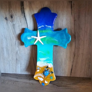 May include: A wooden cross with a blue, green, and yellow resin design depicting a beach scene with a starfish, a sea turtle, and seashells.