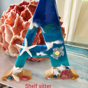 May include: A blue, white, and green resin letter A with a beach scene. The letter is decorated with seashells, starfish, and a turtle. The letter is labeled "Shelf sitter".