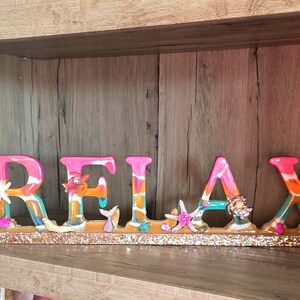 May include: A decorative sign spelling "RELAX" in colorful, stylized letters. The letters are pink, orange, and blue, with seashell and starfish embellishments. The base is covered in gold glitter.