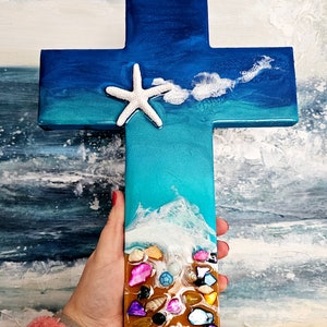 May include: A wooden cross with a blue and turquoise resin background, depicting a beach scene with a white starfish and seashells.