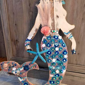 May include: Wooden mermaid wall art with a distressed white finish. The mermaid's tail and body are adorned with blue and pink embellishments, seashells, and a turquoise starfish. A pink feather and seashell accent the head.