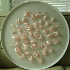 May include: A white circular plate with a decorative design of pink paper cutouts. The cutouts are arranged in a random pattern and resemble a cluster of abstract shapes.