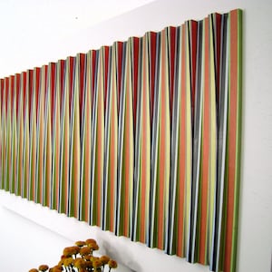 May include: Abstract wall art featuring vertical stripes in shades of red, orange, yellow, green, black, and white. The artwork is mounted on a white rectangular frame and hangs on a white wall. The piece has a textured, dimensional appearance.