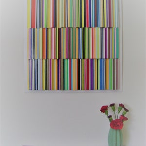 May include: A colourful abstract wall art piece made of vertical wooden strips in various colours, including red, yellow, green, blue, and purple. The art is mounted on a white wall above a wooden shelf with a vase of red carnations.