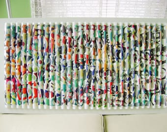 May include: A white wall art piece with a colorful abstract design. The design is made up of many small, rolled-up pieces of paper, each with a different pattern. The paper is arranged in a vertical pattern, creating a sense of movement and depth.