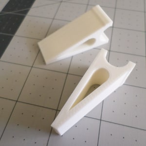 May include: Two white plastic clips with a rounded opening in the center. The clips are designed to hold items in place.