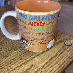 May include: A ceramic mug with an orange exterior and interior, featuring a white handle. The mug displays the text TWO GUN MICKEY, OUR HERO, MICKEY LASSOS HOG TIES THE BAD GUY IN THIS ROOTIN' TOOTIN' 1934 WESTERN ADVENTURE, alongside illustrations of Mickey Mouse.