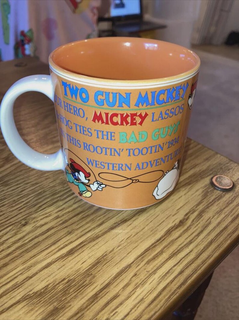 May include: A ceramic mug with an orange exterior and interior, featuring a white handle. The mug displays the text TWO GUN MICKEY, OUR HERO, MICKEY LASSOS HOG TIES THE BAD GUY IN THIS ROOTIN' TOOTIN' 1934 WESTERN ADVENTURE, alongside illustrations of Mickey Mouse.