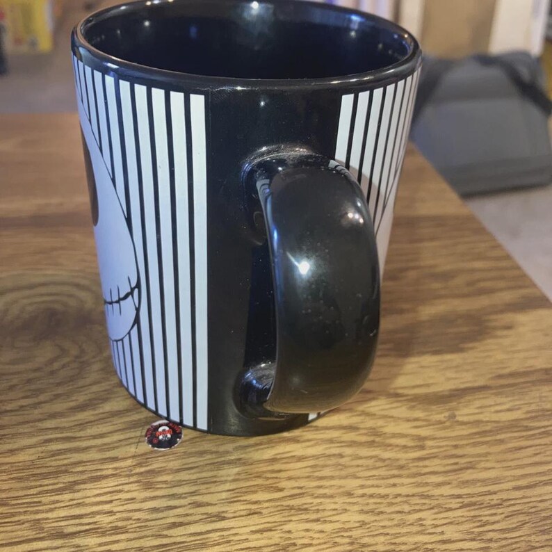 May include: Black ceramic mug with a white and black striped pattern and a cartoon skull face design. The mug has a black handle and is sitting on a wooden surface.