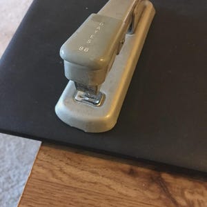 May include: A vintage gray stapler with the text "BATES 88" printed on the top. The stapler is made of metal and sits on a black surface. The stapler is a classic office supply.