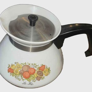 May include: White enamel tea kettle with a black handle and lid knob. The kettle features a floral design with the words "Le Thé" in script. The lid is stainless steel. The kettle is designed for stovetop use.