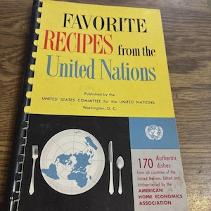 May include: A spiral-bound cookbook titled Favorite Recipes from the United Nations, published by the United States Committee for the United Nations. The yellow cover features blue and red text, a graphic of a plate with a map, and cutlery.