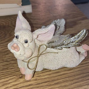 May include: A whimsical figurine of a pig with angel wings. The pig is light beige with pink accents on the ears, snout, and feet. It has a gold-colored bow tied around its neck and gold-tipped wings. The figurine is a decorative item.