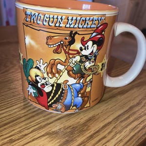 May include: A ceramic mug with a light orange interior and exterior featuring a graphic of Two Gun Mickey. The illustration depicts Mickey Mouse as a cowboy on a horse with a lasso, alongside another character, set against a desert background.