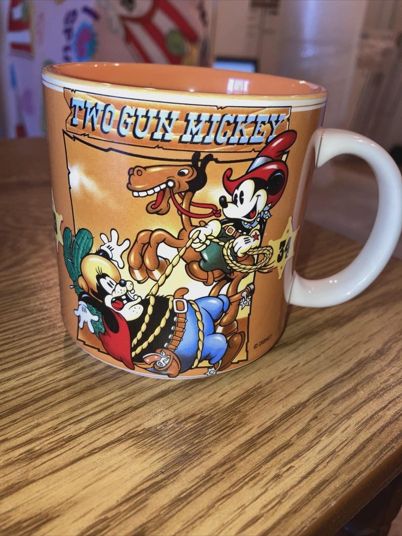 May include: A ceramic mug with a light orange interior and exterior featuring a graphic of Two Gun Mickey. The illustration depicts Mickey Mouse as a cowboy on a horse with a lasso, alongside another character, set against a desert background.