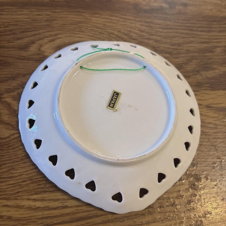 May include: White ceramic decorative plate with heart-shaped cutouts around the edge. A green string is attached for hanging. The plate has a sticker that reads "JAPAN".