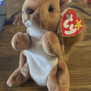May include: A plush brown squirrel toy with a white belly and a red heart-shaped tag. The squirrel has small black eyes, whiskers, and a fluffy tail. The tag reads "Beanie Original Baby".