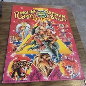 May include: A vintage Ringling Bros. and Barnum & Bailey Circus poster. The red poster features illustrations of circus performers, animals, and the text "Ringling Bros. Barnum & Bailey Circus". The poster includes images of a lion, a clown, and a man riding an alligator.