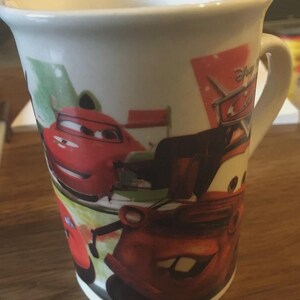 May include: White ceramic mug featuring characters from the Disney Cars film. The mug has a curved handle and is decorated with images of Lightning McQueen and Mater. The mug is on a wooden surface.