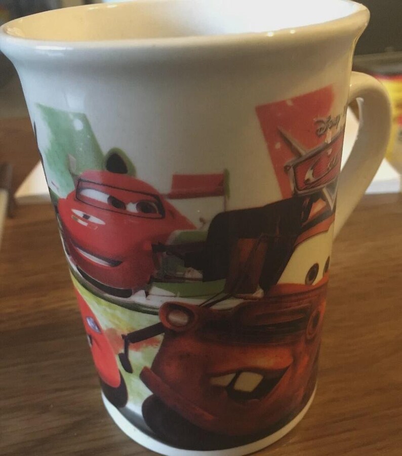 May include: White ceramic mug featuring characters from the Disney Cars film. The mug has a curved handle and is decorated with images of Lightning McQueen and Mater. The mug is on a wooden surface.