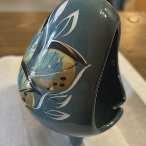 May include: A teal ceramic napkin holder with a butterfly and leaf design. The holder features a curved shape with a small base and a slot for napkins. The butterfly wings are blue and yellow with white accents.