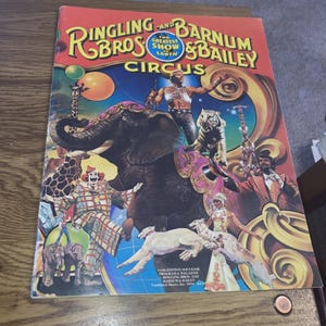 Peut inclure: Un ancien programme et magazine souvenir du cirque Ringling Bros. and Barnum & Bailey. La couverture présente une illustration colorée d'artistes de cirque, d'animaux et le texte "The Greatest Show on Earth". La couverture est rouge avec des lettres dorées.