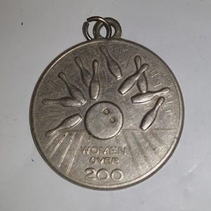 May include: A silver-colored bowling medal with a circular design. The medal features a bowling ball in the center, surrounded by bowling pins. The text "WOMEN OVER 200" is inscribed below the ball.
